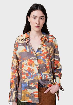 Donna in camicia a maniche lunghe con motivo astratto arancione, blu, grigio e beige, che indossa pantaloni marroni con una mano in tasca.