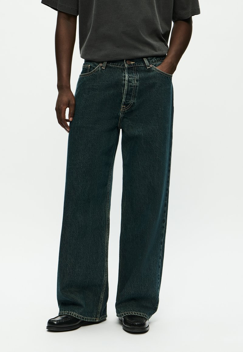 Persoon met donkere wijde jeans, zwarte schoenen en een donkergrijze t-shirt met één hand in de broekzak, tegen een effen witte achtergrond.