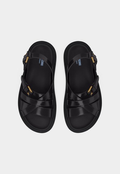 Ζευγάρι μαύρων δερμάτινων σανδαλιών με λουριά που διασταυρώνονται και χρυσά αγκράφες, φωτογραφημένα από ψηλά σε φωτεινό φόντο.