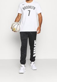 Valkoinen Nike t-paita, jossa on mustalla painettu "BROOKLYN" ja numero 7, yhdistetty mustiin urheiluhousuihin, joissa on valkoinen korostus. Pitelee koripalloa.