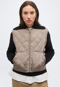 Vrouw die een beige doorgestikt vest draagt over een zwarte gebreide trui, witte blouse en beige broek, staand met handen in de zakken van het vest.