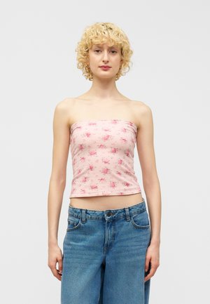 Personne aux cheveux blonds bouclés portant un haut bustier rose à fleurs et un jean taille haute bleu, debout devant un fond uni.