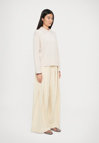 Maglione beige chiaro con colletto, abbinato a pantaloni larghi color crema. L'insieme presenta una consistenza morbida e una vestibilità rilassata e comoda. Scarpe a righe.