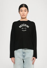 Mladá žena v čiernom tričku s dlhým rukávom s nápisom "Moschino 86-23 Jeans", skombinovaným so svetlými rifľovými džínsy s vysokým pásom, stojí na bielom pozadí.