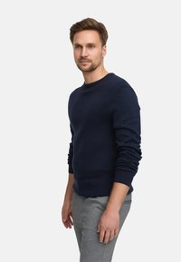 Mann med lys skjegg iført en marineblå teksturert genser og grå bukser, som står mot en ensfarget lys bakgrunn.