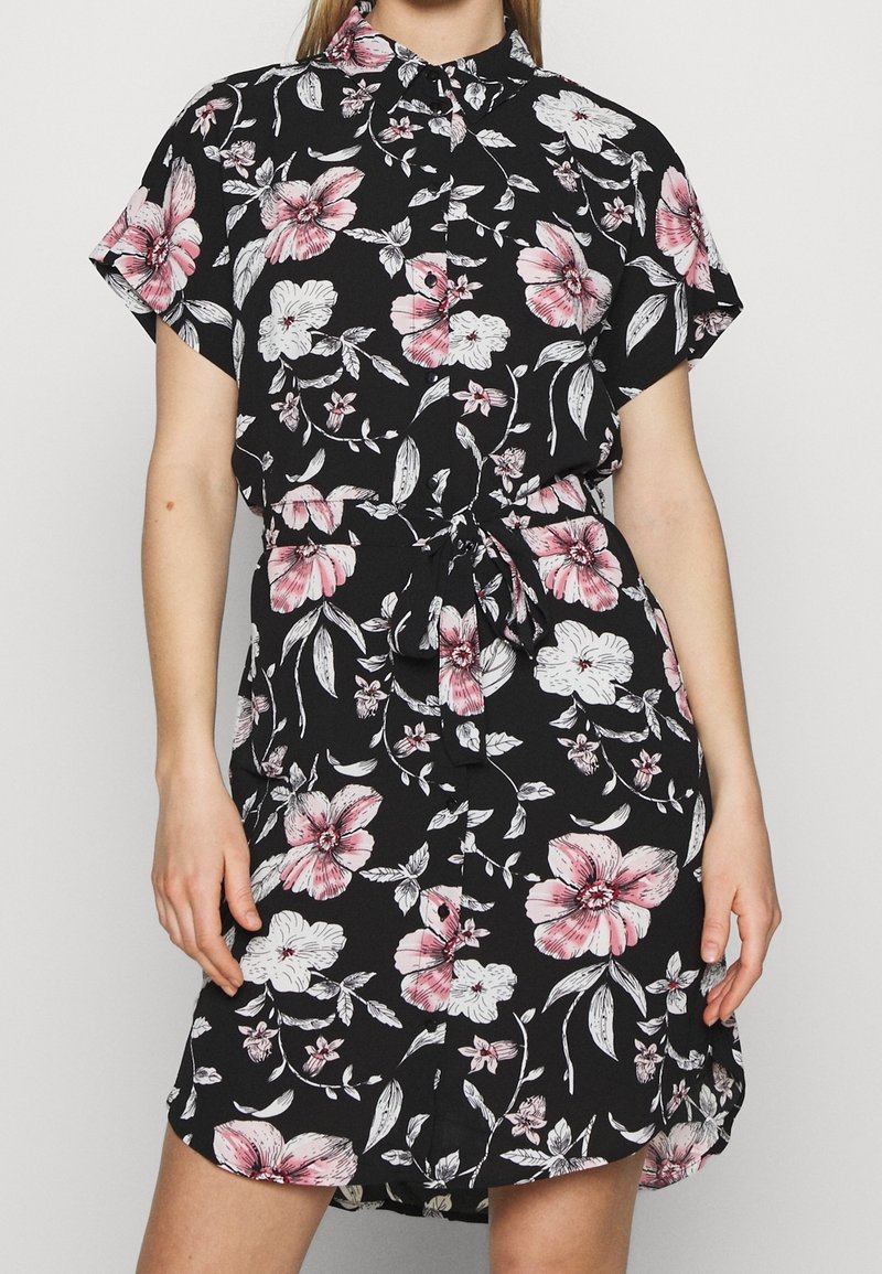 Femme portant une robe noire à manches courtes avec un imprimé floral rose et blanc et une ceinture nouée à la taille, debout devant un fond uni.