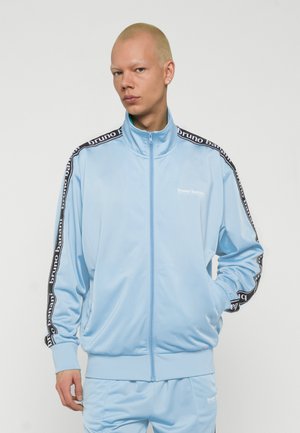 Hombre con cabello rubio corto que lleva chaqueta y pantalones de chándal azul claro de Bruno Banani, de pie con una mano en el bolsillo contra un fondo liso.