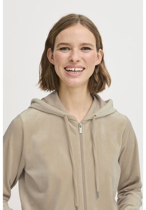 Ung kvinne med kort brunt hår som smiler, iført en beige hettegenser med glidelås og snorer mot en enkel lys bakgrunn.