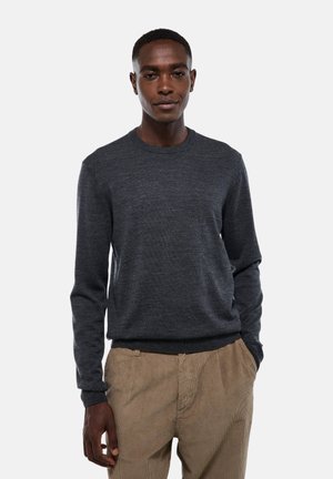 Homme portant un pull à manches longues gris foncé et un pantalon en velours côtelé beige, se tenant avec une main dans sa poche devant un fond uni.
