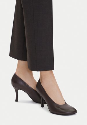 Kobieta nosząca ciemnobrązowe skórzane szpilki na lekko rozkloszowanych obcasach oraz czarne, przycięte, dopasowane spodnie.