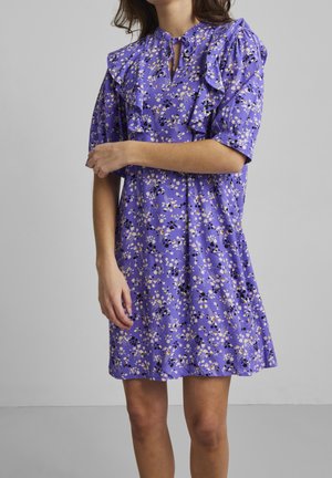 Robe florale violette avec des manches froncées et un décolleté en trou de serrure. Présente des motifs floraux blancs, jaunes et noirs sur un tissu doux.