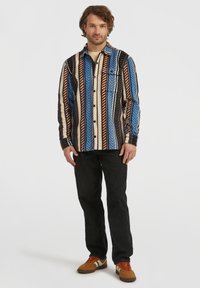 Hombre de pie con una camisa de botones de rayas verticales multicolores, pantalones negros y zapatillas marrones con rayas blancas y cordones naranjas.
