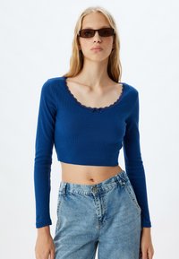 Sininen pitkähihainen crop-top, jossa on ribbitekstuuri ja aallotettu kaula-aukko. Yhdistetty korkeavyötäröisten haalistuneiden farkkujen kanssa.