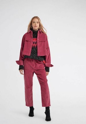 Donna in piedi che indossa una giacca e pantaloni di velluto a coste rosa scuro, un maglione nero con motivo rosso e scarpe col tacco nere.