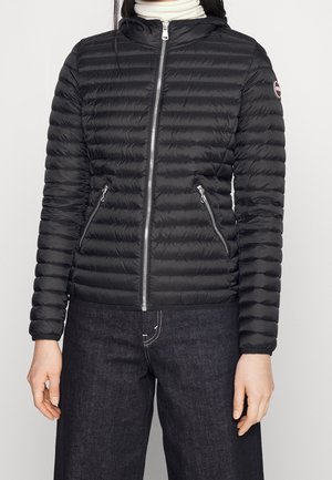 Žena v černé prošívané bundě na zip se dvěma zipovými kapsami a tmavých džínách, stojící před jednobarevným pozadím.