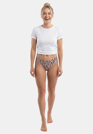 Femme souriante aux cheveux blonds en chignon, portant un t-shirt blanc court et un sous-vêtement rose à imprimé léopard, marchant pieds nus sur un fond uni.