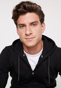 Zwarte zip-up hoodie met trekkoord en witte accenten, gedragen boven een wit T-shirt. Glad textuur van de stof, casual pasvorm, gecentreerde close-up.