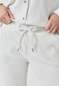 Witte geribde loungewear set met een knoopsluiting bovenstuk en broek met drawstring en zilveren metalen tips. De stof heeft een zachte, gestructureerde uitstraling.