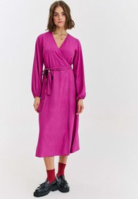 Różowa sukienka kopertowa z długimi rękawami, o fakturowanym wzorze, z paskiem w talii, zestawiona z czerwonymi skarpetkami i czarnymi platformami.