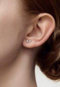 Srebrne kolczyki w kształcie liści noszone są w płatku ucha. Ich design charakteryzuje się prostymi liniami i gładkim metalicznym wykończeniem, które pięknie kontrastuje z jasną karnacją.
