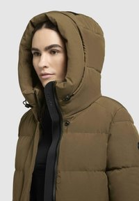 Puffjakke i olivengrøn med en stor, aftagelig hætte, der har en lynlåsfortil, sorte accenter og en quiltet tekstur.