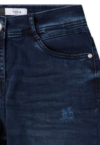 Tummansininen denimkangashousut, joissa on sileä pinta, etutasku ja hopeinen nappi. Huomattava haalistuma kankaassa.