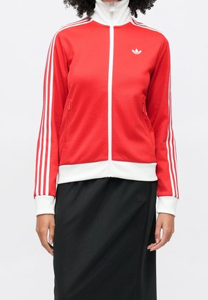 Personne portant une veste Adidas rouge et blanche zippée avec des bandes blanches sur les manches et une jupe noire.