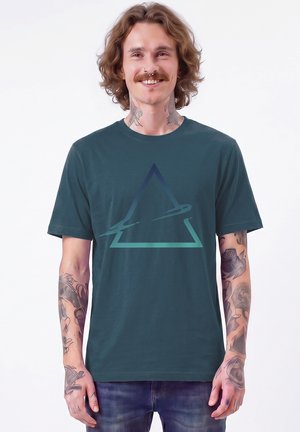 Jonge man met krullend haar en tatoeages op nek en armen glimlacht, draagt een teal T-shirt met een gestileerd driehoekig ontwerp.