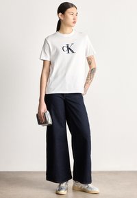 Valge puuvillane T-särk tumesinise "ck" logoga, sobitatud tumedate laiade pükstega. Aksessuaarideks on metalliktoonis kott ja hallid tossud.