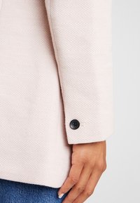 Jasnoróżowy płaszcz wykonany z teksturowanej tkaniny z wzorem w jodełkę. Posiada czarny guzik na mankiecie rękawa.