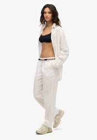 Witte linnen shirt, zwarte gebreide bralette, witte losvallende broek met zwarte logoband, en beige sneakers met textuurdetails.