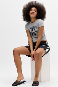 Mladić s afro frizurom, obučen u sivi majicu s grafikom i crne Adidas šortse, sjedi na bijelom bloku s crnim japankama.