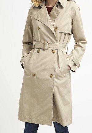 Beige double-breasted trench coat with belt, buttoned cuffs, flap shoulder detail, worn over dark pants with hands in pockets.