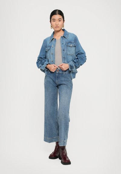 Ženska, ki stoji, oblečena v modro jeans jakno, ujemajoče se široke hlače, bež top, zlato uhane v obliki obroča in burgundske gležnjarje na belem ozadju.