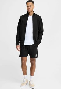Giacca nera con zip sopra una t-shirt bianca, abbinata a pantaloni corti neri. Calzature: sneakers beige con lacci bianchi. Stile semplice e atletico.