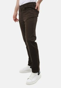 Bruine slim-fit broek met een gladde textuur, voorzien van zijzakken en een knoopsluiting, gecombineerd met witte sneakers met zwarte accenten.