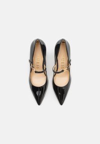 Talons hauts en cuir verni noir avec des bouts pointus, une bride de cheville et un marquage doré à l'intérieur. Texture lisse et finition brillante.