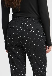 Zwarte broek met een rekbare tailleband, voorzien van een wit hartjespatroon, twee achterzakken en een gladde, lichte stoftextuur.