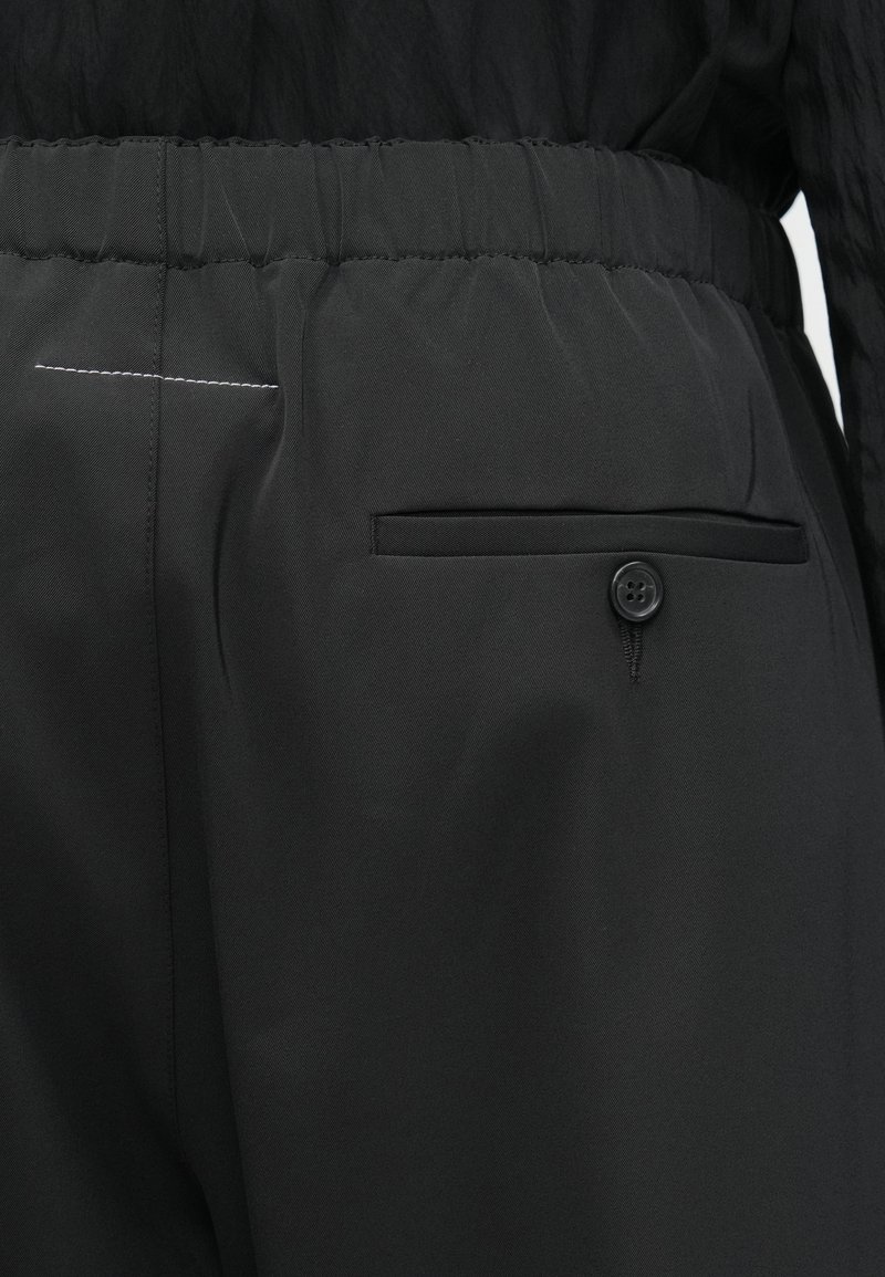 Nærbillede af sort bukselinning med elastisk ryg, knaplukket brystlomme og hvid stikningsdetalje nær venstre side.