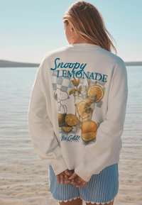 Sweatshirt crème avec un graphique de Snoopy buvant une limonade, accompagné de citrons et d'une boisson. Le design comprend du texte teal et un fond à carreaux.