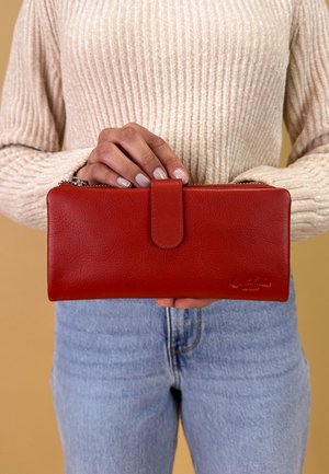 Red leather wallet with a textured surface, zip closure, and a tab fastener. Held by a hand against a beige background, paired with light jeans.