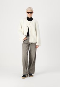 Veste blanche en fourrure avec des boutons ronds, associée à un jean gris à jambes larges et des baskets noires. Le mannequin porte des lunettes de soleil sombres. Fond minimaliste.