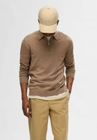 Beige Sweatshirt mit Reißverschluss und Polokragen, gestrickte Textur und langen Ärmeln, kombiniert mit hellbeigen Chinos und einer passenden Schirmmütze.