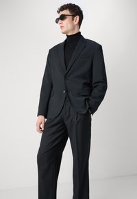 Completo nero su misura con giacca a un bottone e pantaloni a gamba dritta. Presenta un dolcevita sotto e occhiali da sole scuri. Tessuto liscio.