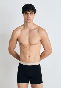 Schwarze Boxer-Briefs mit einem beigen Bund, der mit "HUGO" beschriftet ist. Der Stoff wirkt glatt und die Passform ist körpernah.