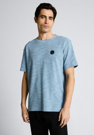 Junger Mann mit dunklen Haaren, der ein hellblaues, strukturiertes, kurzärmliges Hemd mit einem kleinen schwarzen runden Logo auf der Brust trägt.