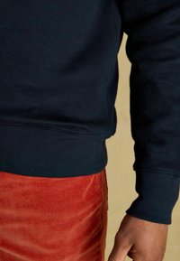 Tummansininen collegepaita, jossa on ribbikuvat hihansuissa ja helmassa, yhdistettynä teksturoidun punaisen samettihousun kanssa. Lähikuva kankaan yksityiskohdista ja istuvuudesta.