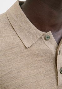 Artimas vaizdas į beige megztą polo marškinėlį su mygtukais tamsios odos žmogaus kaklo ir viršutinės krūtinės srityje.