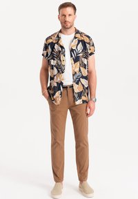 Camicia blu navy a maniche corte con un motivo di foglie tropicali, abbinata a pantaloni beige e scarpe chiare. T-shirt bianca visibile sotto.