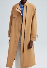 Beżowy płaszcz o prostym kroju, z zamkiem błyskawicznym z przodu, guzikiem na mankietach i boczną kieszenią. Nosi go w zestawieniu z niebieską koszulą i jasnymi spodniami.